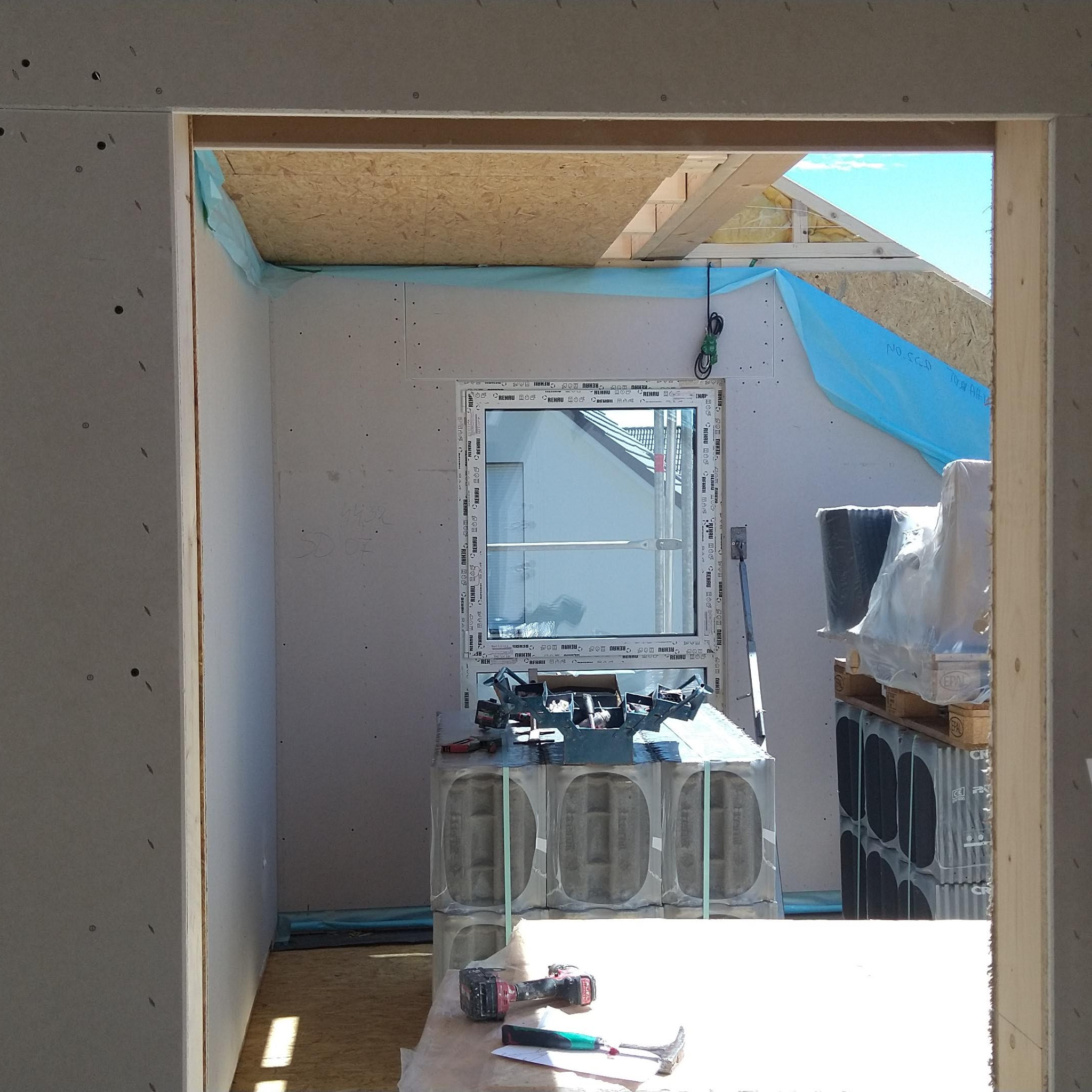 Wnętrze w trakcie prac z płytami gipsowo-kartonowymi, widoczne narzędzia i materiały budowlane. Okno z widokiem na zewnątrz. Niebieska folia zabezpieczająca.