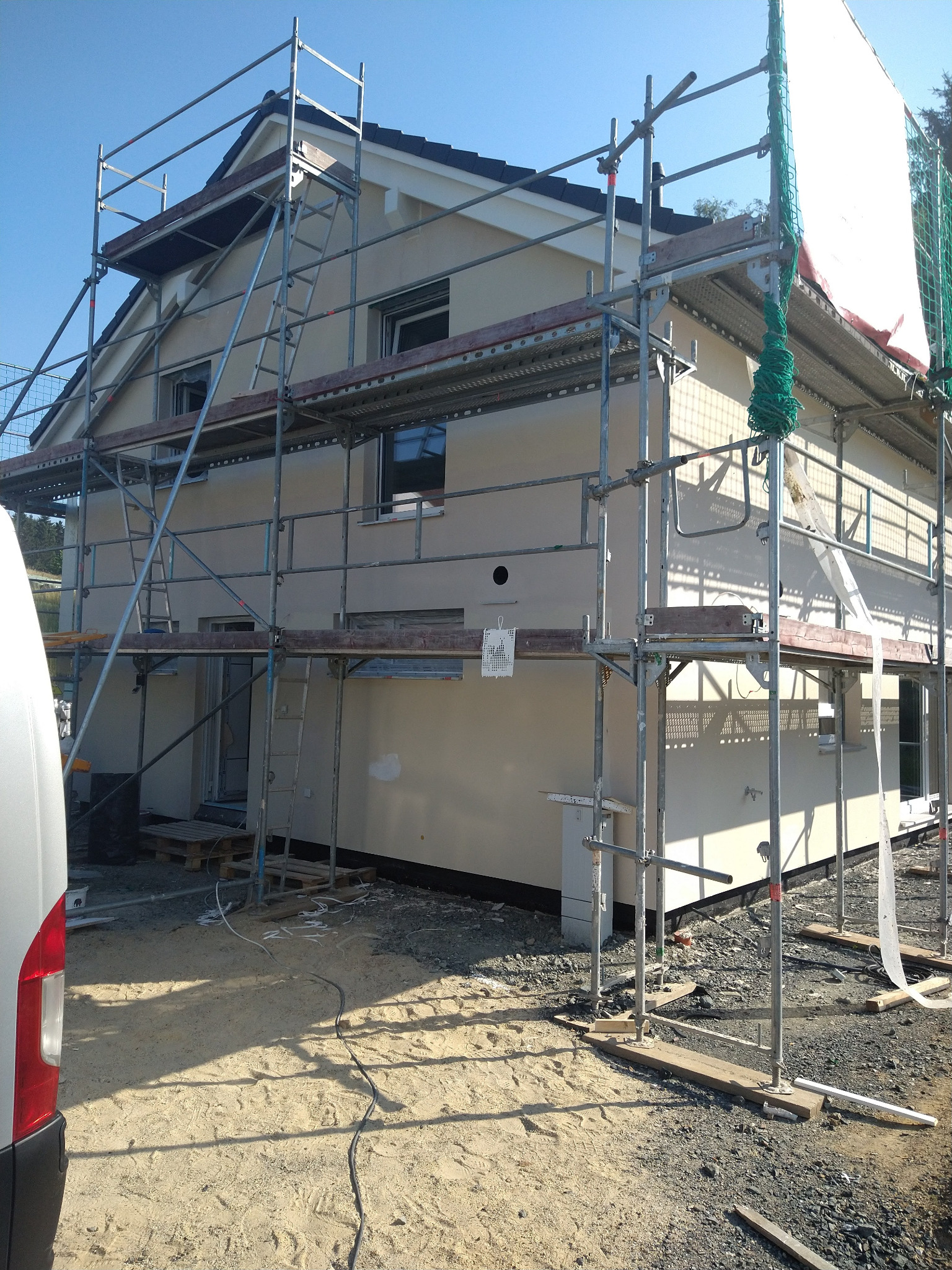 Dom w trakcie prac elewacyjnych, otoczony rusztowaniem. Widoczne świeżo nałożone warstwy tynku. Częściowo zasłonięty siatką ochronną. W tle błękitne niebo.