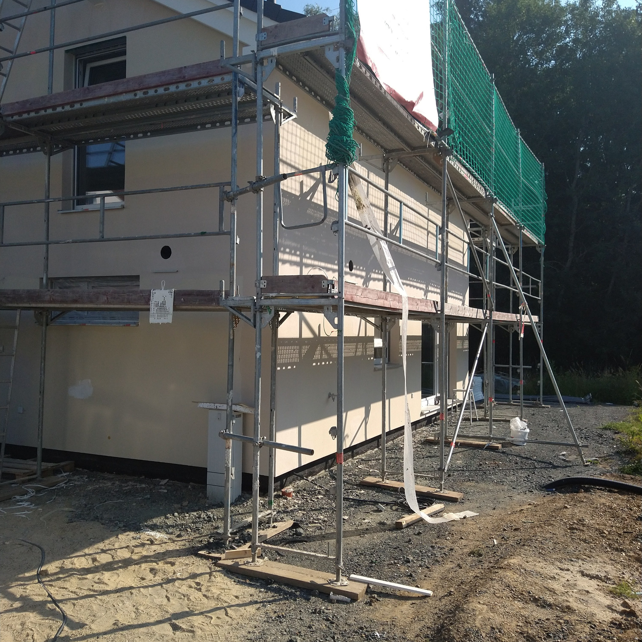 Elewacja domu w trakcie prac wykończeniowych, widoczne rusztowania i siatka ochronna. Świeżo otynkowana ściana w jasnym odcieniu. Ujęcie z dołu, w tle drzewa.