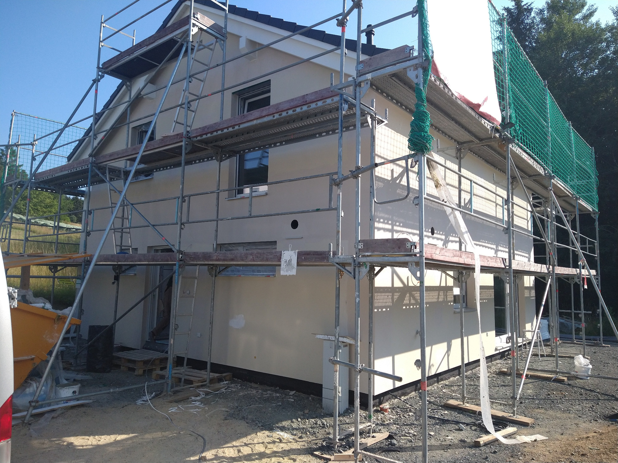 Dom jednorodzinny w trakcie prac wykończeniowych elewacji, otoczony rusztowaniem. Widoczne otwory wentylacyjne w ścianie i siatka zabezpieczająca na dachu.