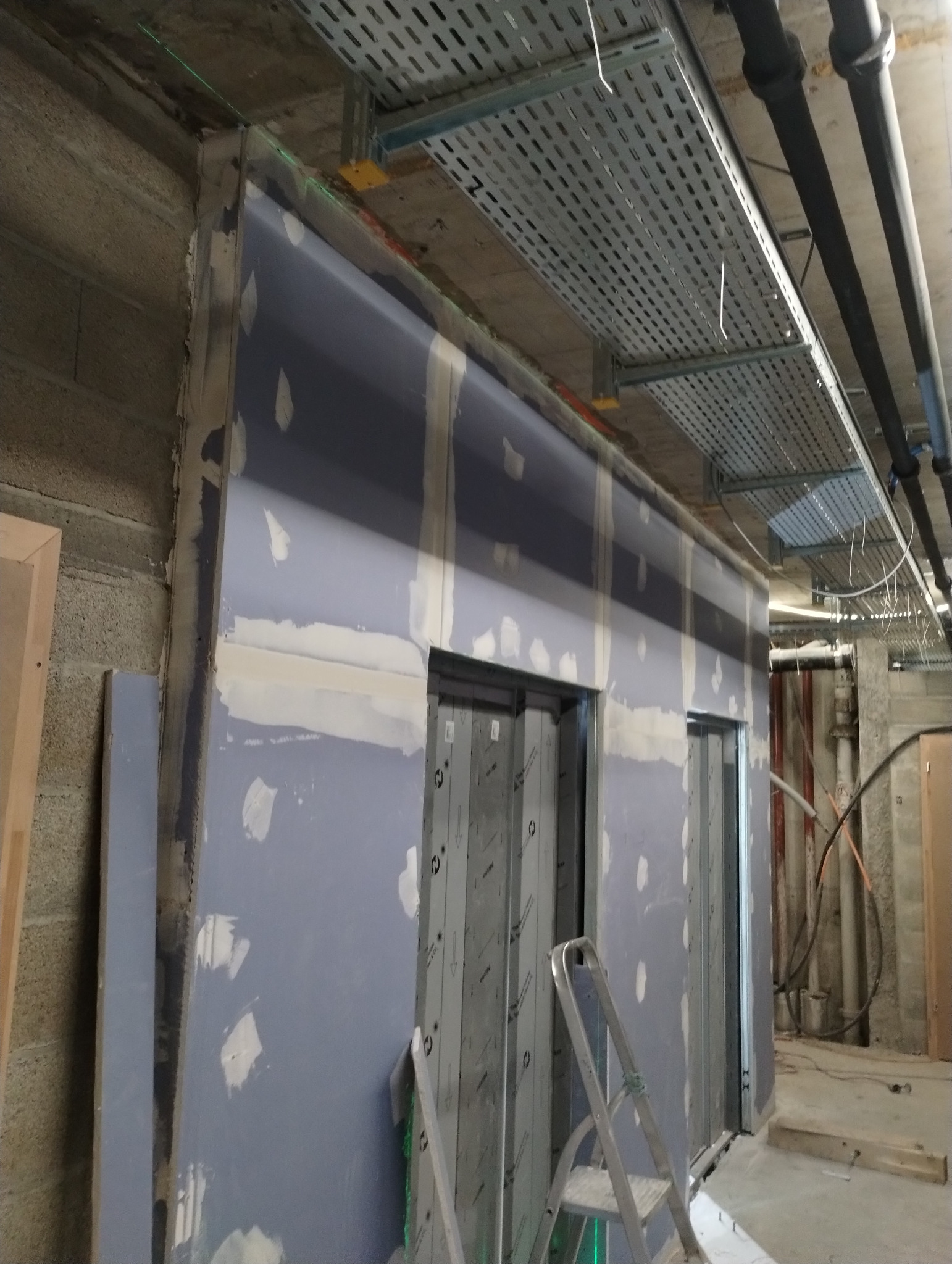 Montaż ścianki działowej z płyty gipsowo-kartonowej w przestrzeni biurowej. Widoczne profile, płyty, szpachlowanie łączeń, drabina. Surowy stan, instalacje pod sufitem.