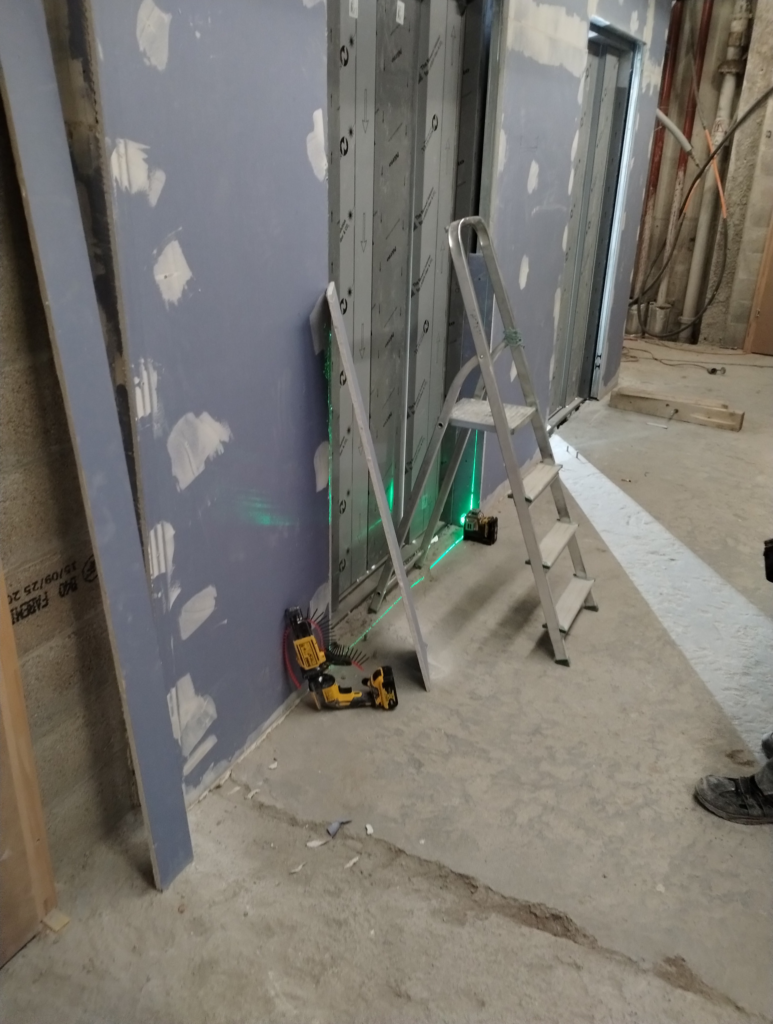 Montaż stelaża pod płyty gipsowo-kartonowe z użyciem poziomicy laserowej. Widoczna drabina i narzędzia. Ściany przygotowane do dalszych prac wykończeniowych.