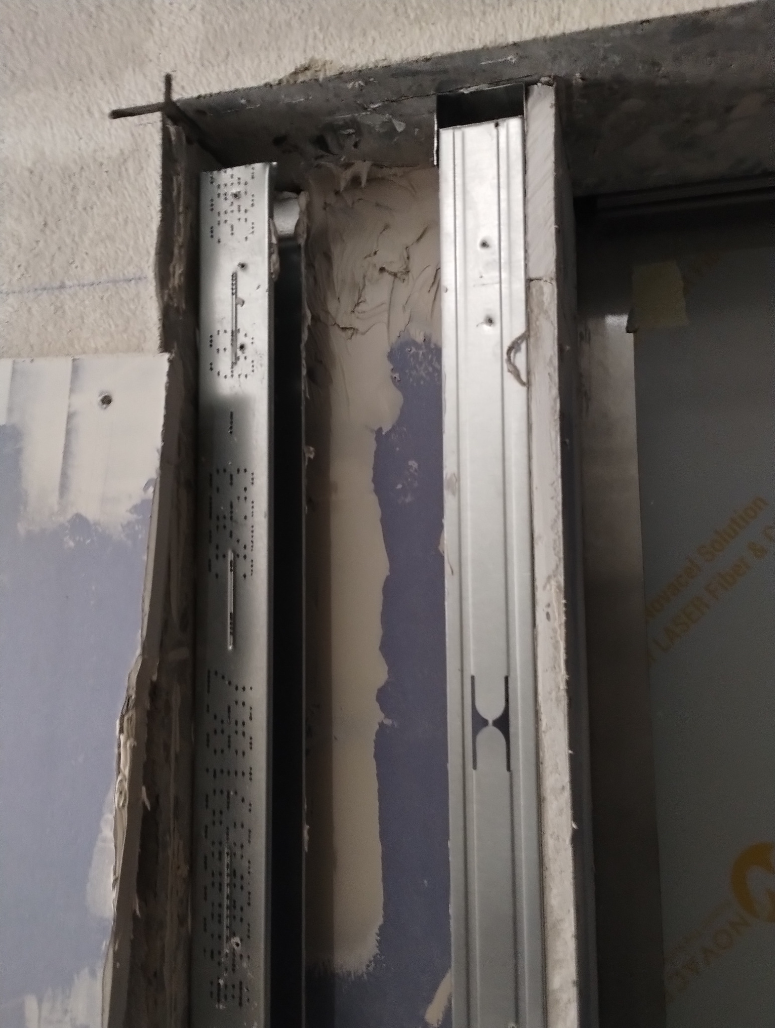 Montaż metalowych profili do płyt karton-gips na ścianie. Widoczne elementy konstrukcyjne i fragment płyty. Prace wykończeniowe w trakcie realizacji.