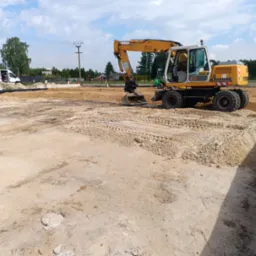 Żółta koparka Liebherr 900 na placu budowy, przygotowująca teren pod fundamenty, widoczne ślady opon na piasku, w tle budynek i drzewa pod błękitnym niebem.