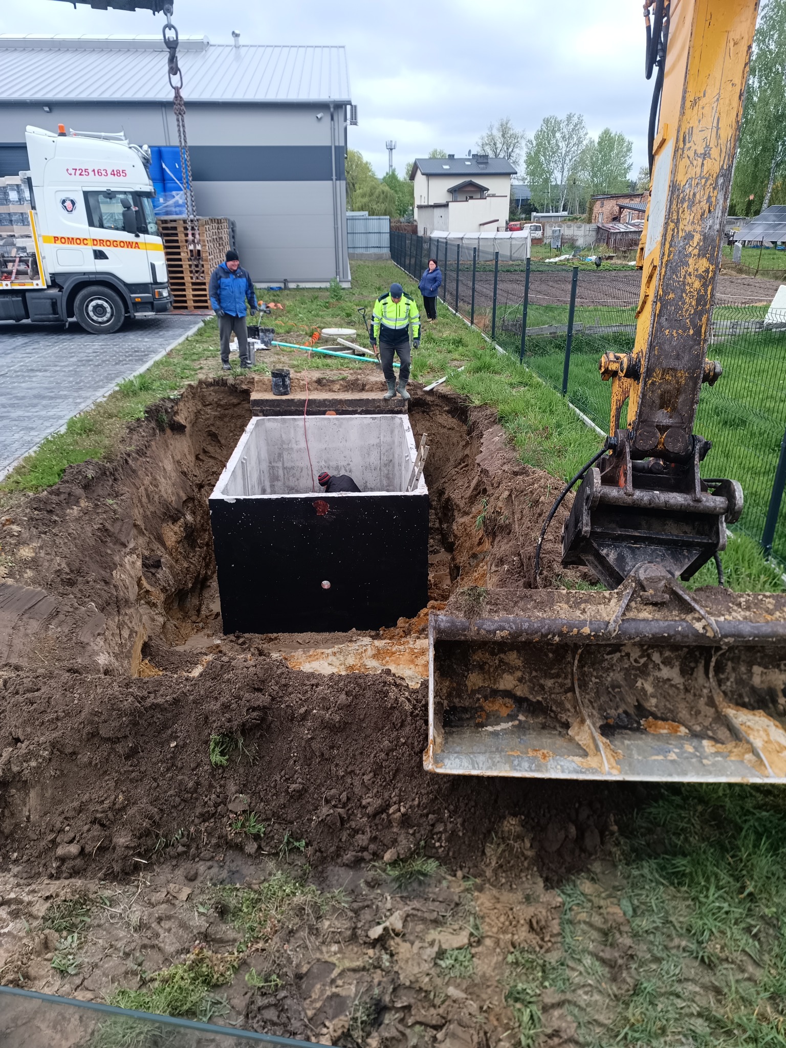 Betonowy zbiornik w wykopie, pracownicy i koparka na placu budowy, widoczny transport materiałów na paletach