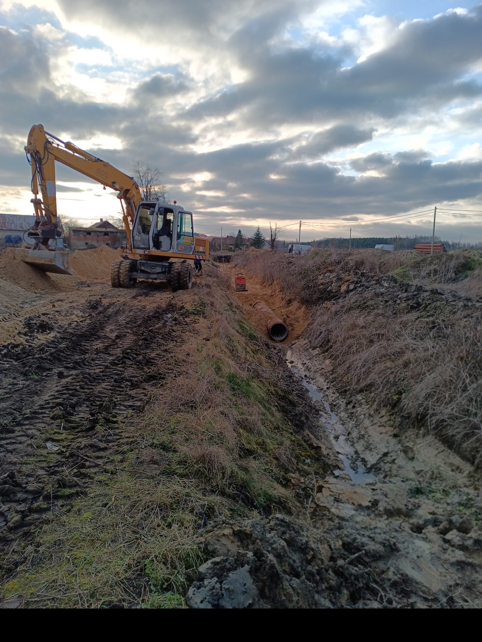 Żółta koparka Liebherr przy wykopie z ułożoną rurą kanalizacyjną w ziemi, widoczne ślady opon i błoto, pochmurne niebo w tle.