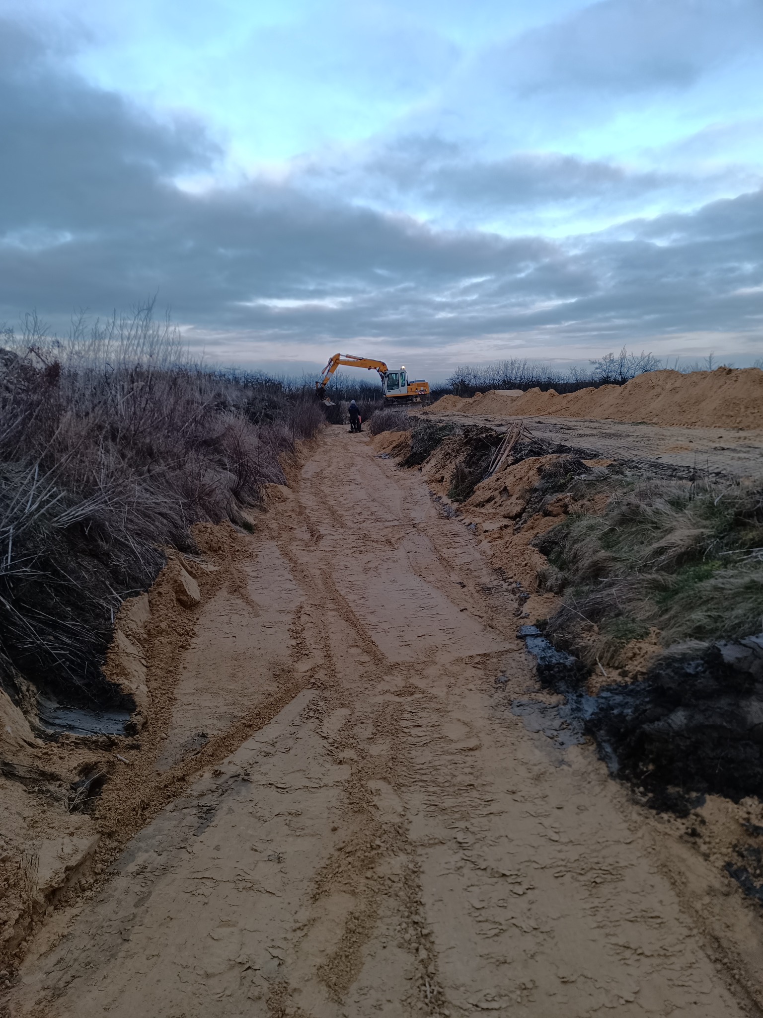 Wykopany rów z widocznymi śladami opon, w tle pracująca koparka na tle pochmurnego nieba.