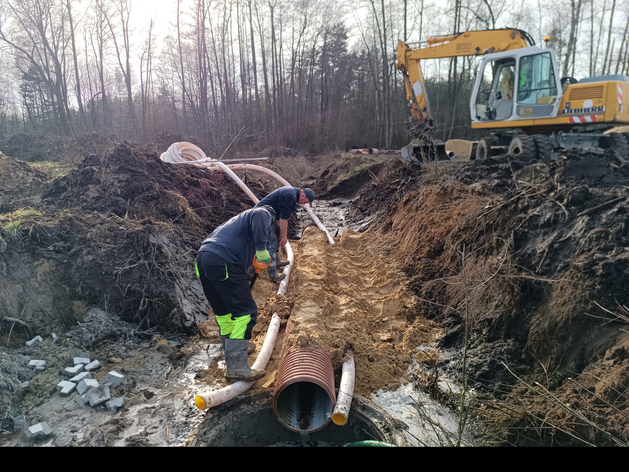 Dwóch pracowników układa rury drenażowe w wykopie, z widoczną koparką Liebherr w tle i stertami ziemi, na terenie z drzewami bez liści.