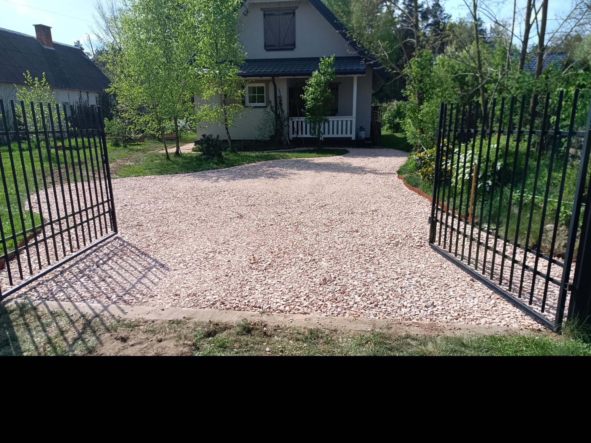 Podjazd do domu wysypany jasnym żwirem, otwarty czarny metalowy płot, widok na dom z jasną elewacją i zielenią w tle.