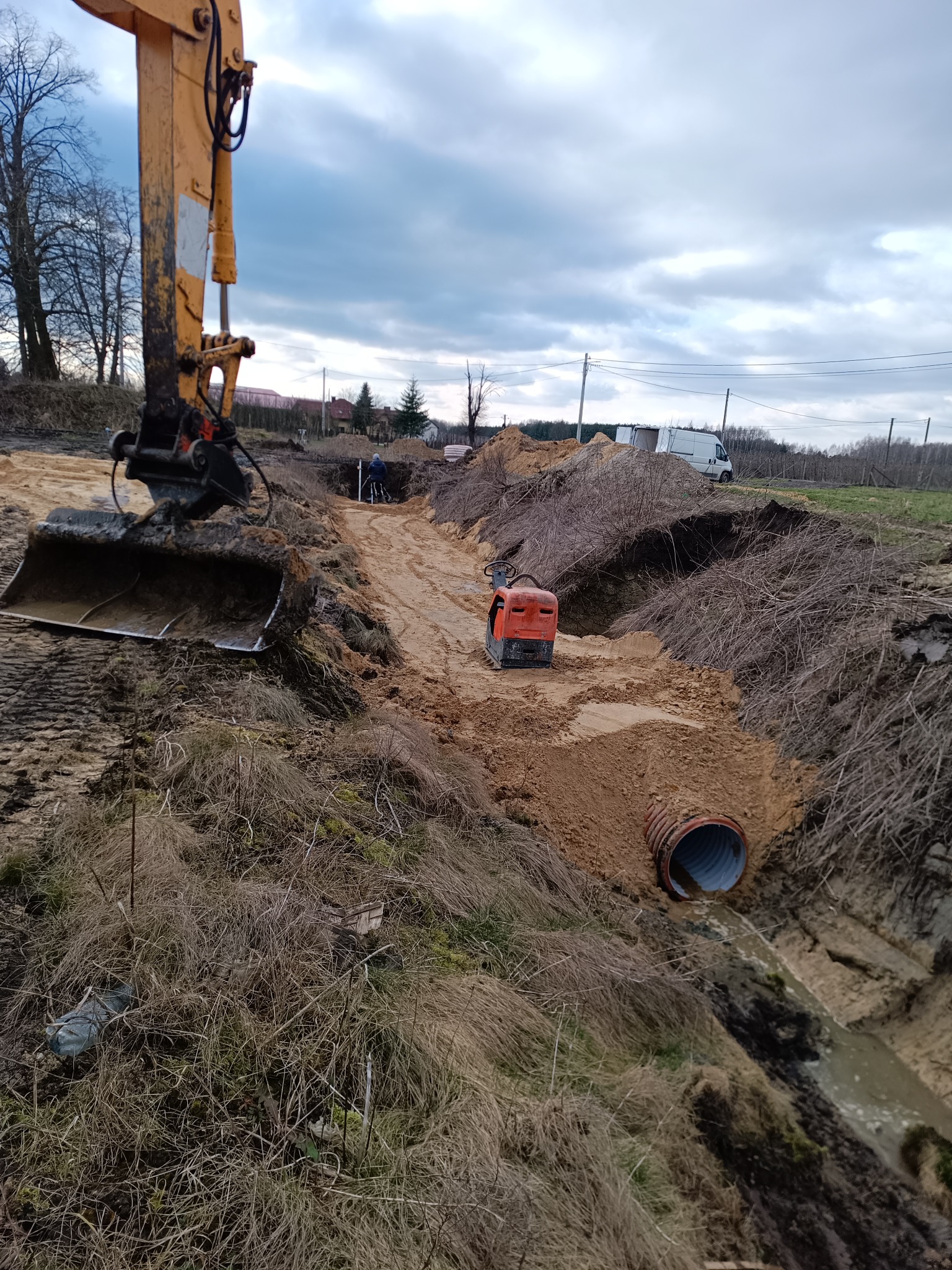 Żółta koparka przy wykopie z pomarańczową rurą i zagęszczarką, widoczny pracownik w oddali, pochmurne niebo.