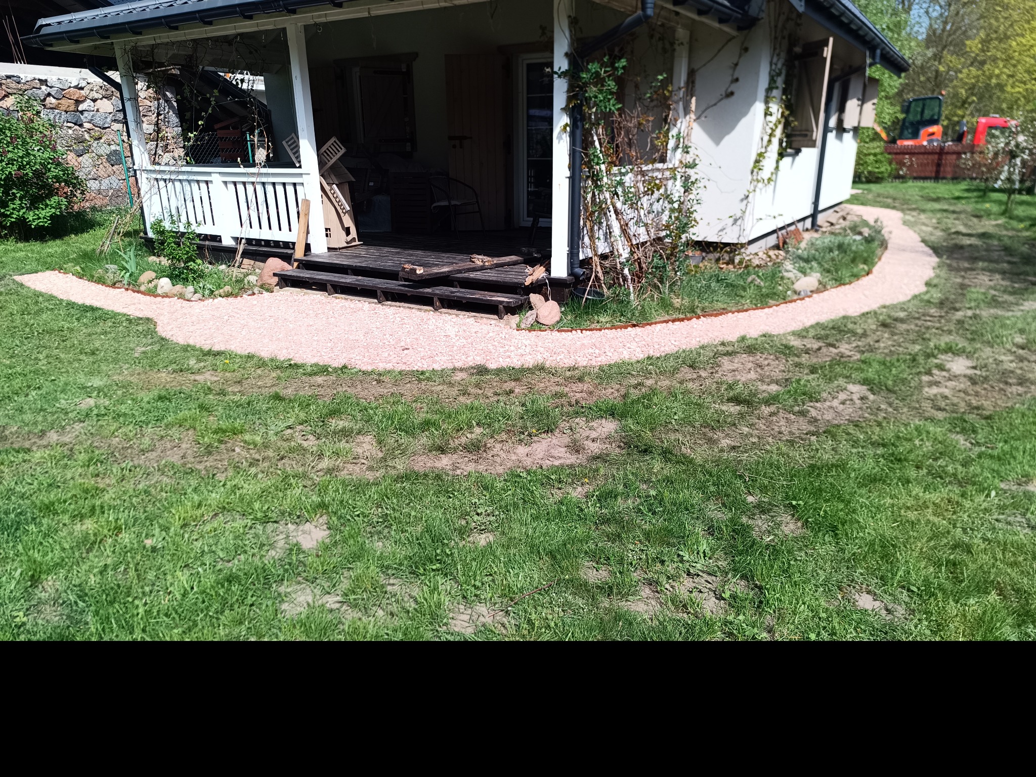 Kruszywowa ścieżka ogrodowa prowadząca do drewnianego tarasu przy domku, z widocznym w tle małym traktorem.