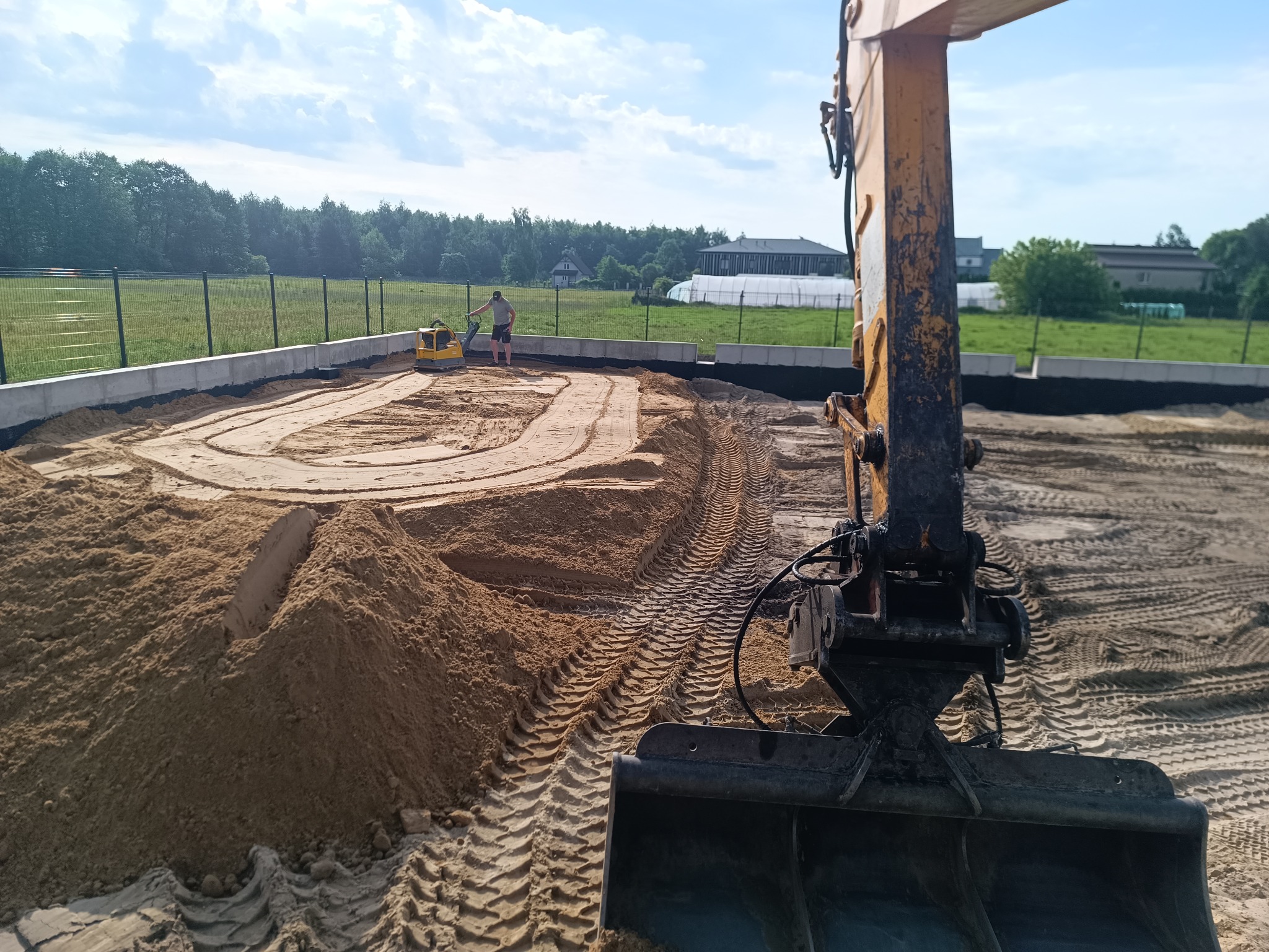 Przygotowanie terenu pod budowę w Białobrzegach: ubijanie piasku zagęszczarką w wykopie z fundamentami, widoczna łyżka koparki i ogrodzenie.
