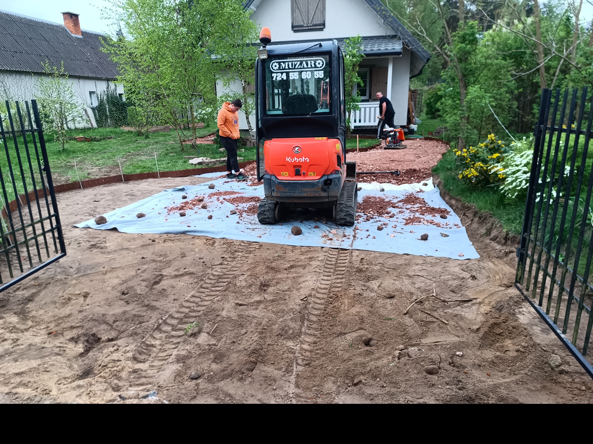 Mini koparka Kubota na posesji, przygotowanie terenu pod utwardzenie, widoczne ślady opon w piasku, pracownicy i zagęszczarka w tle.
