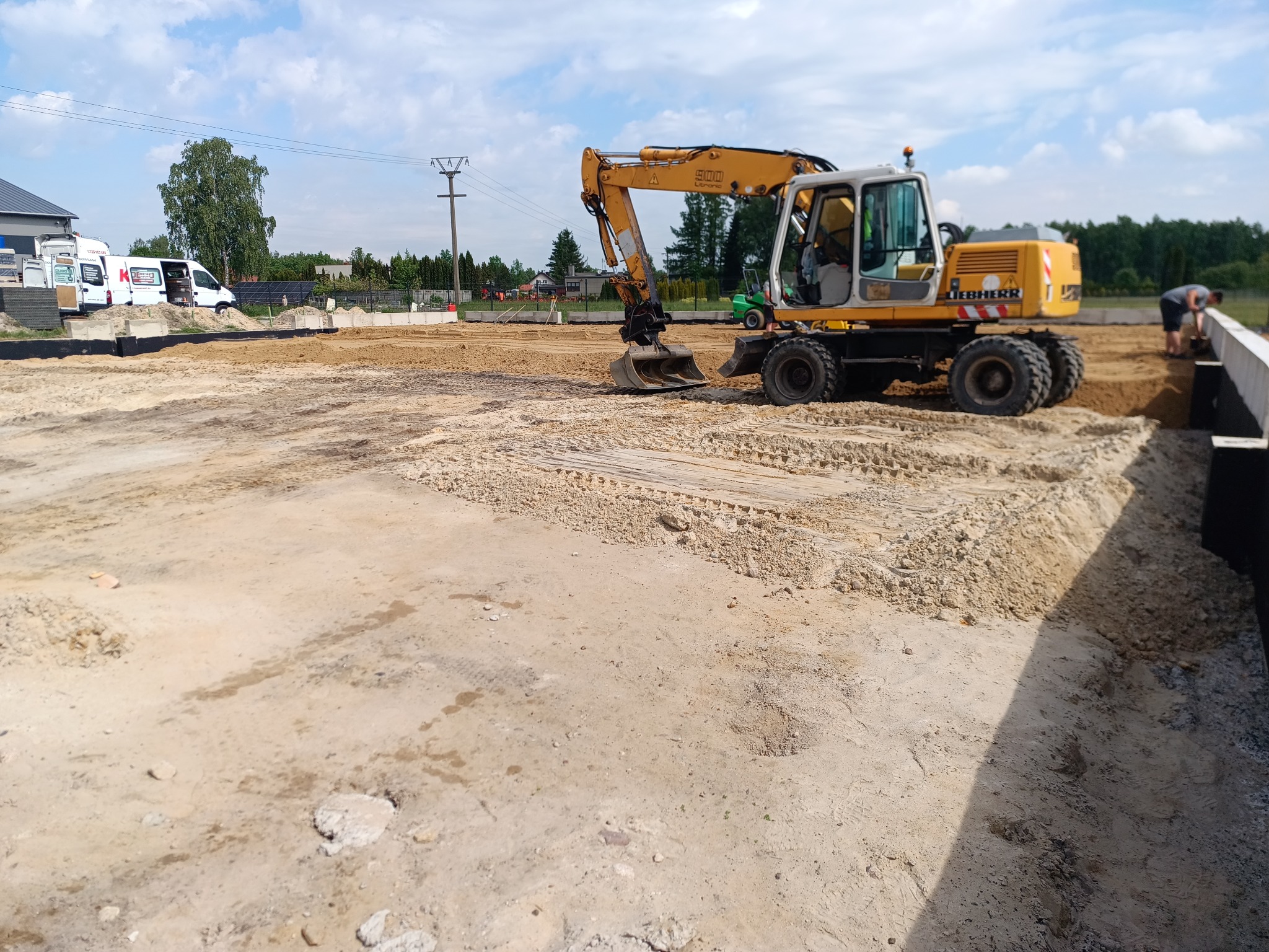 Żółta koparka Liebherr 900 na placu budowy, przygotowująca teren pod fundamenty, widoczne ślady opon na piasku, w tle budynek i drzewa pod błękitnym niebem.