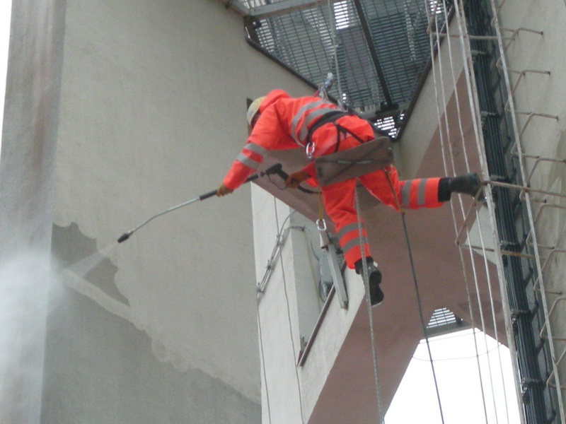 Pracownik w pomarańczowym kombinezonie, zabezpieczony linami, myje elewację budynku wysokociśnieniową myjką. Widoczny fragment elewacji po umyciu, kontrastujący z brudną powierzchnią. W tle...