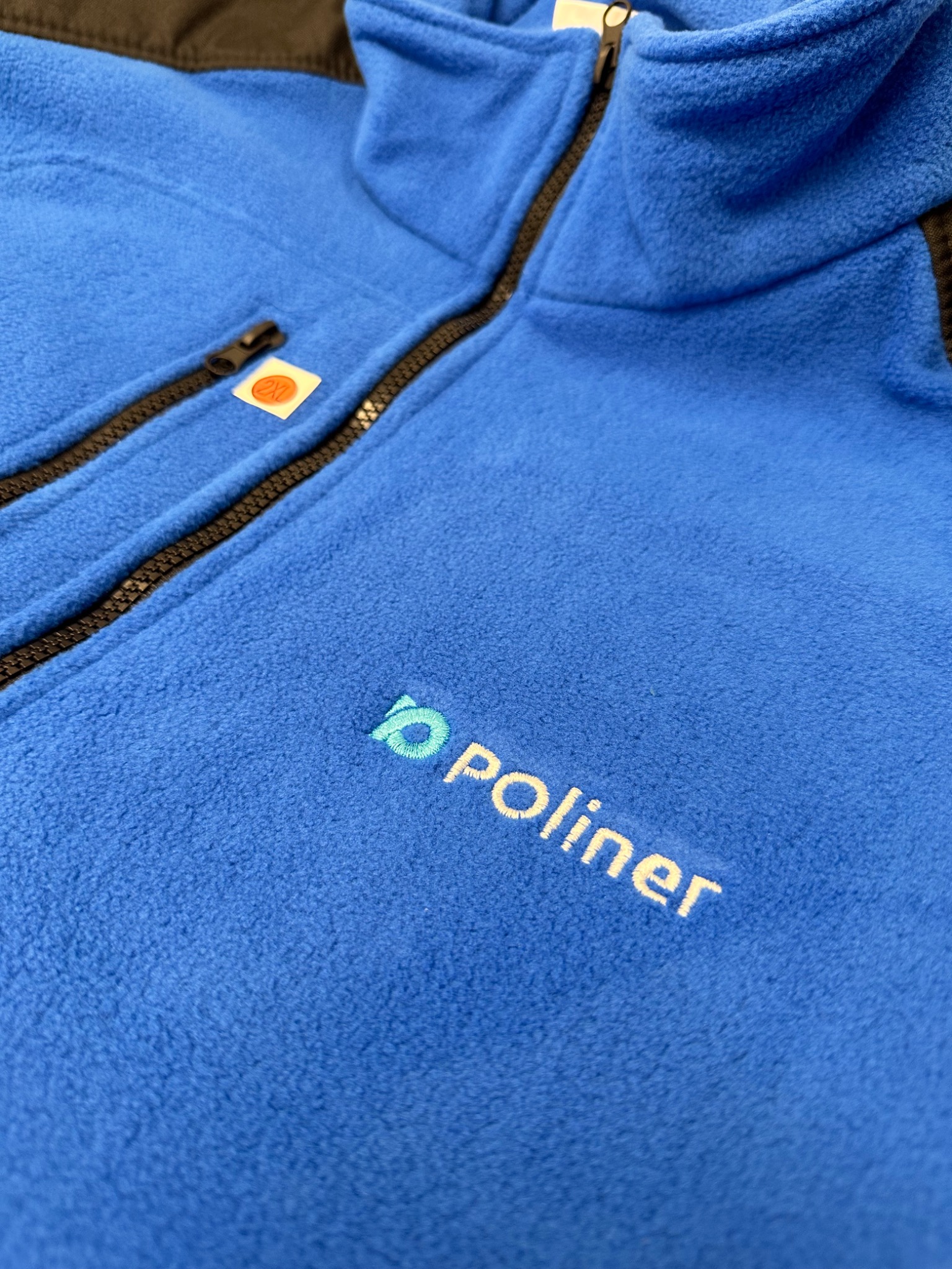 Niebieska bluza polarowa z czarnym zamkiem błyskawicznym i wyhaftowanym logo 'poliner' w kolorze niebiesko-białym, widoczna metka z rozmiarem 2XL.
