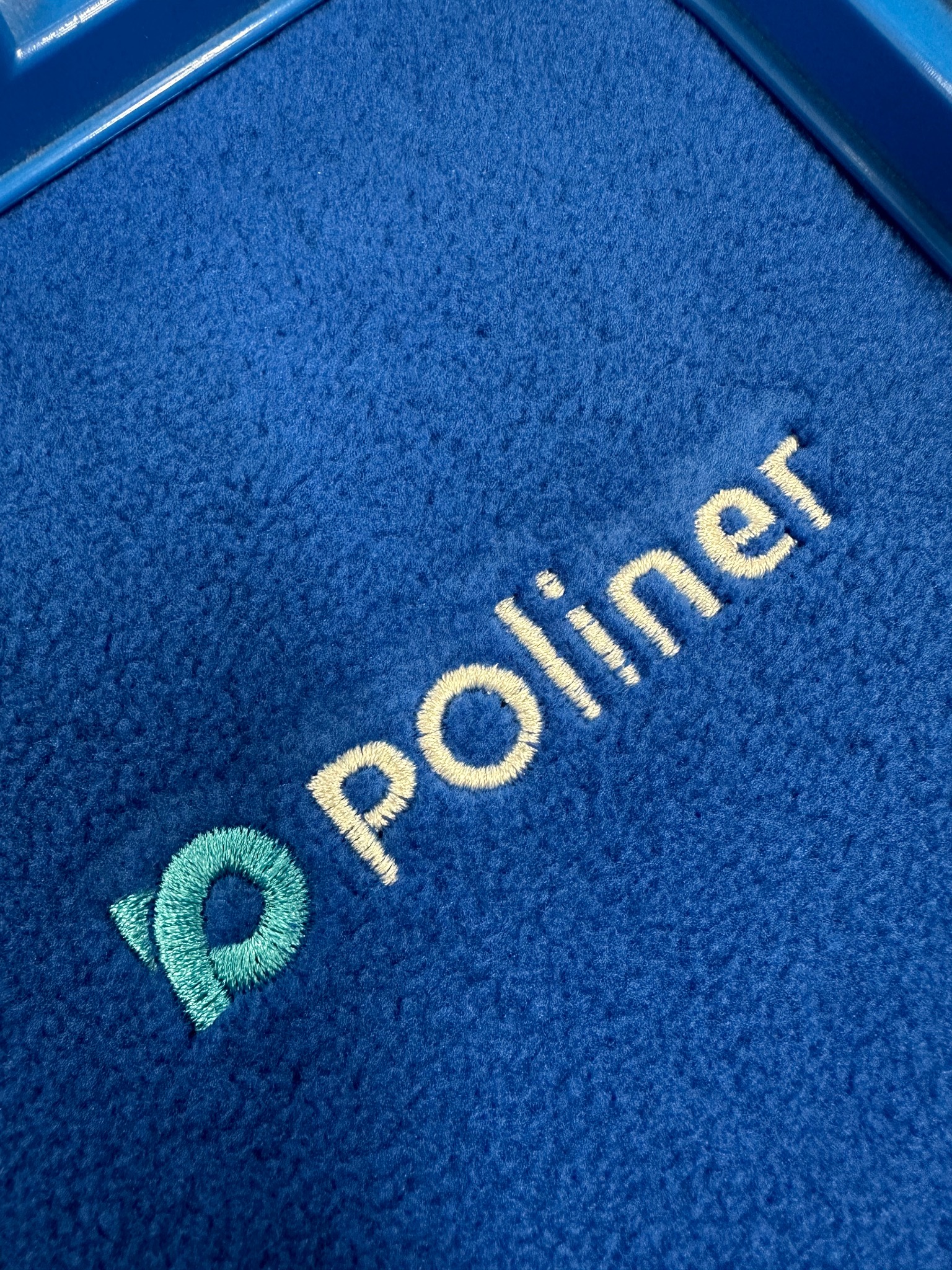 Niebieski materiał frotte z wyhaftowanym logo i napisem 'Poliner' w kolorze turkusowym i kremowym, umieszczony w niebieskiej ramce.