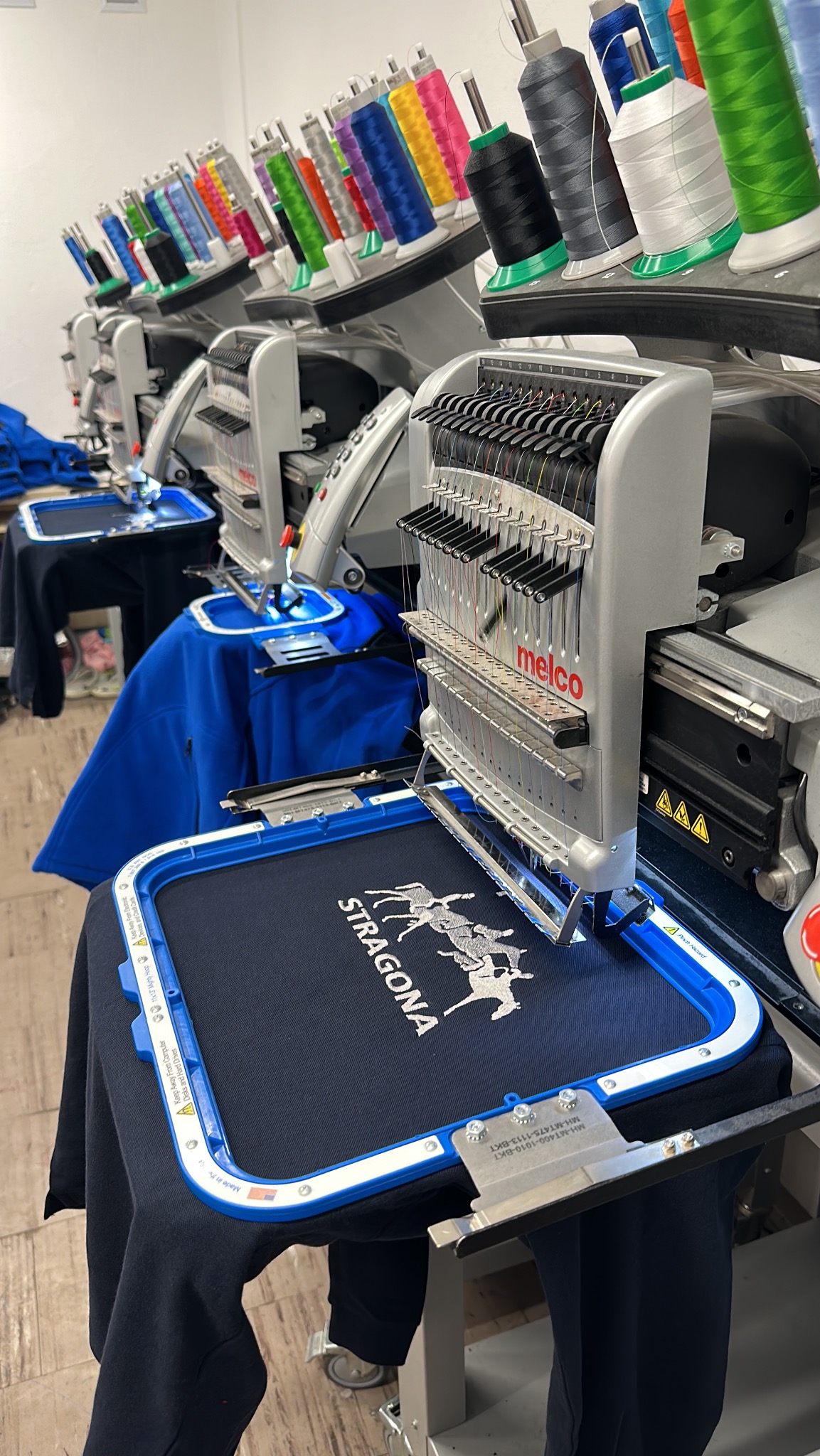 Szereg profesjonalnych maszyn do wyszywania, z nałożonymi kolorowymi nićmi, w trakcie pracy nad wzorem jeźdźców i napisu 'STRAGONA' na granatowej koszulce, umieszczonej w niebieskiej ramce.