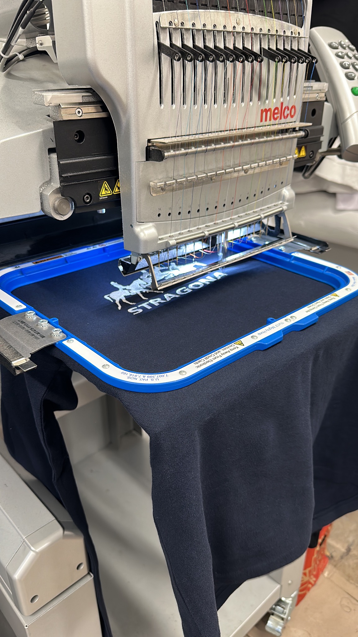Automatyczna maszyna do szycia, w trakcie wykonywania białego haftu przedstawiającego jeźdźca na koniu i napis STRAGONA na granatowej koszulce, umieszczonej w niebieskiej ramce.