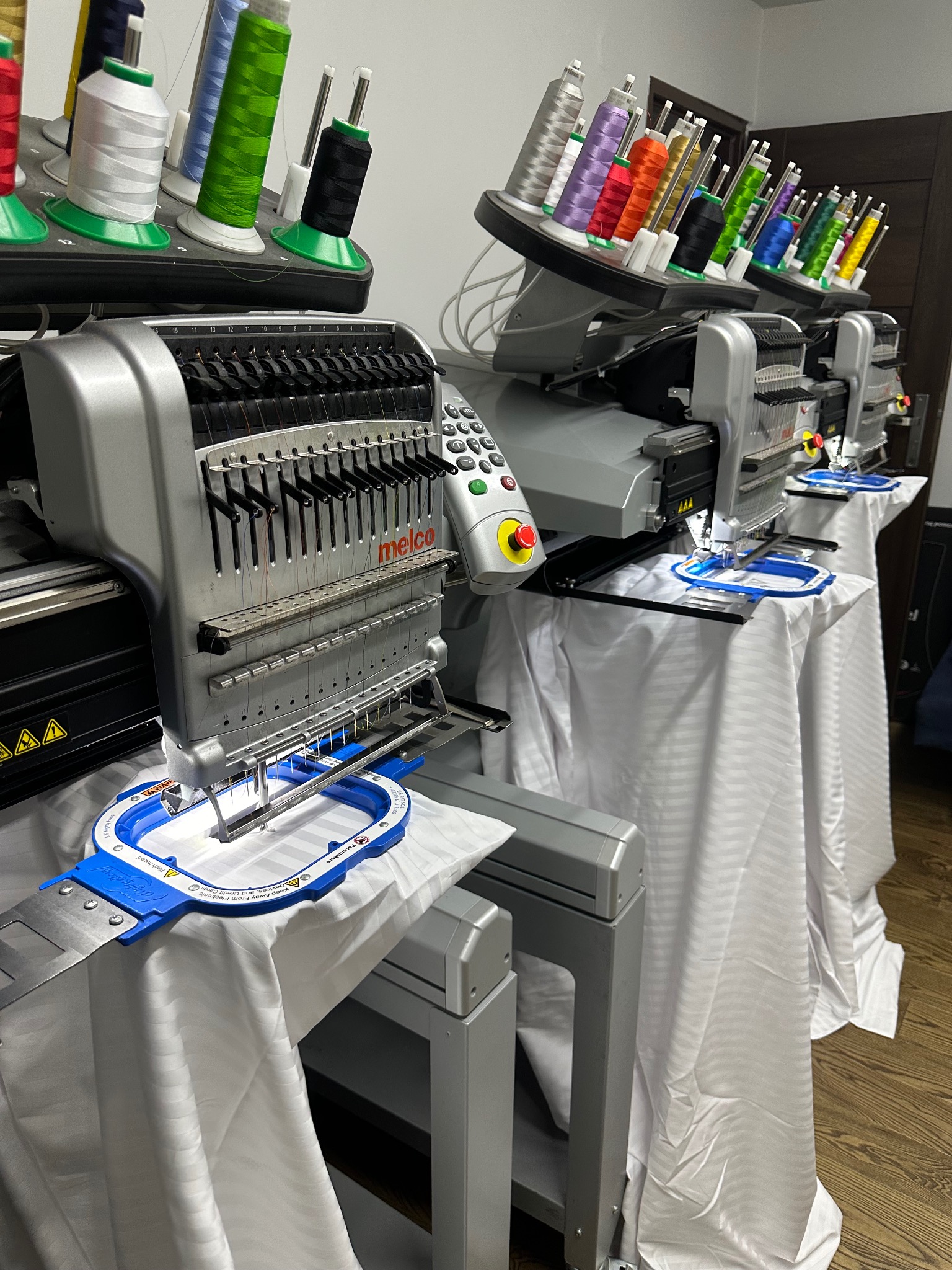 Rząd profesjonalnych maszyn do szycia z kolorowymi nićmi, przygotowanych do pracy nad wzorem na białej tkaninie.