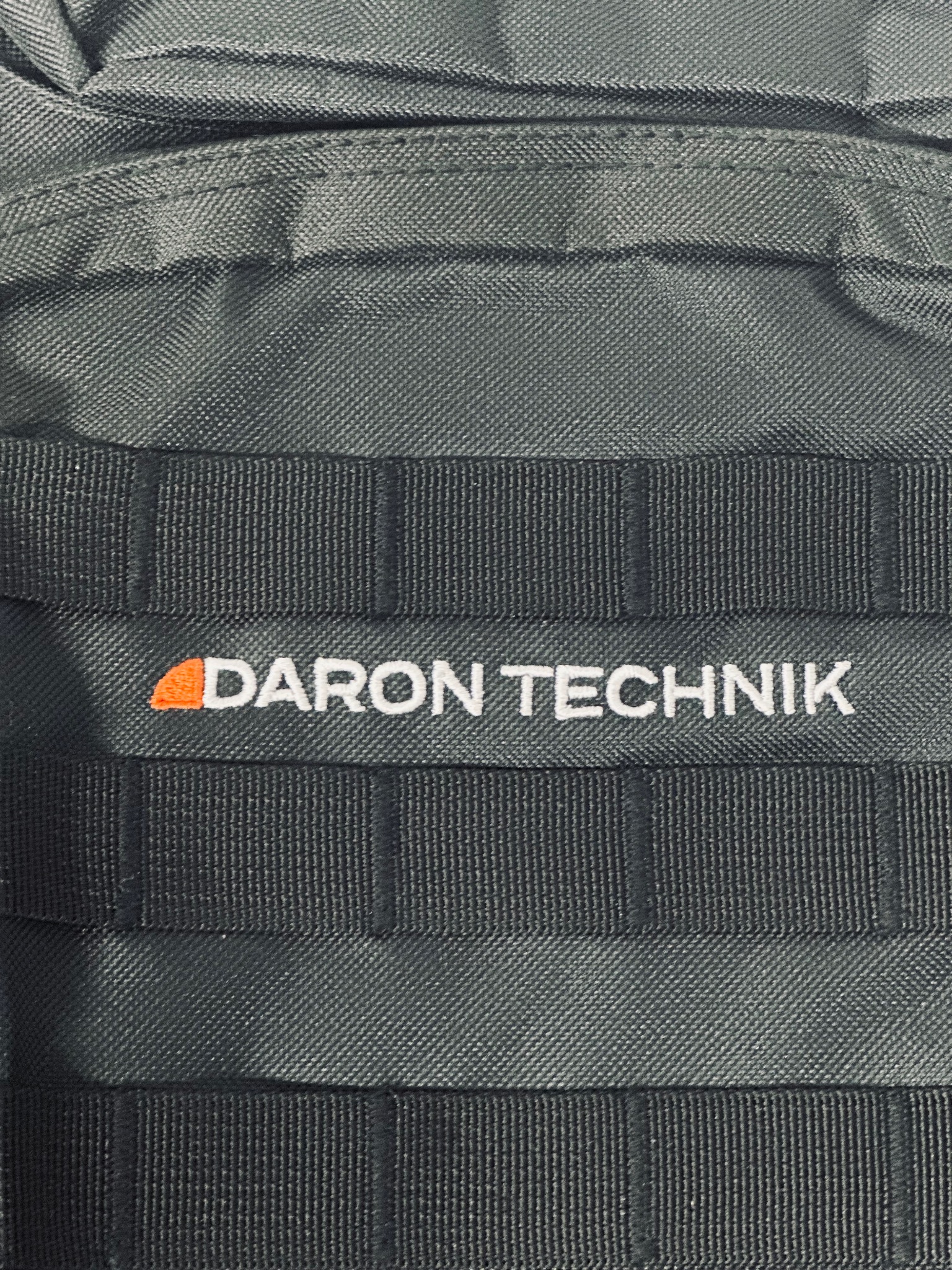 Szczegółowe ujęcie fragmentu plecaka taktycznego z naszytym logo 'DARON TECHNIK' w kolorze pomarańczowo-białym na tle ciemnoszarej tkaniny z systemem mocowania MOLLE.