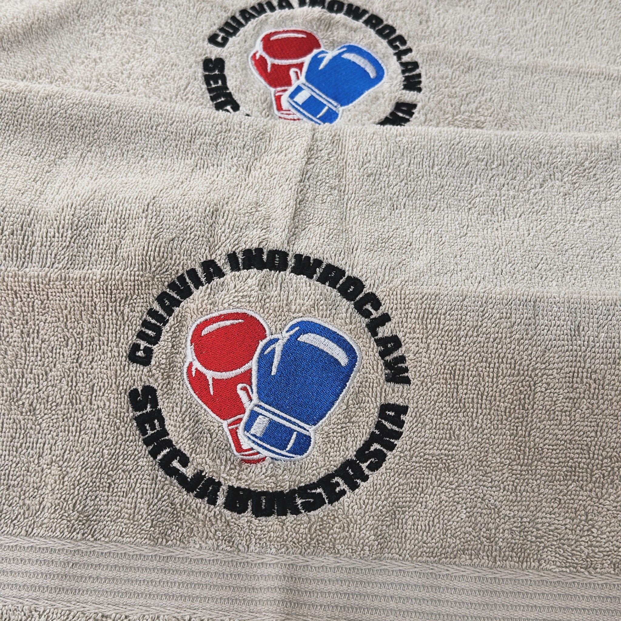 Beżowy ręcznik frotte z wyhaftowanym logo klubu bokserskiego: czerwona i niebieska rękawica bokserska otoczone czarnym napisem 'SEKCJA BOKSERSKA CUIAVIA IKO WROCŁAW'.