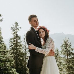 Dreamcatchers Kuba&Ula Jakub Ćwiklewski Fotograf Ełk - Para młoda, mężczyzna w czarnym garniturze i kobieta w koronkowej sukni i wianku z kwiatów, obejmująca się na tle gór i lasu iglastego w słoneczny dzień.