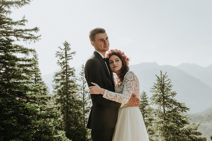 Para młoda, mężczyzna w czarnym garniturze i kobieta w koronkowej sukni i wianku z kwiatów, obejmująca się na tle gór i lasu iglastego w słoneczny dzień.