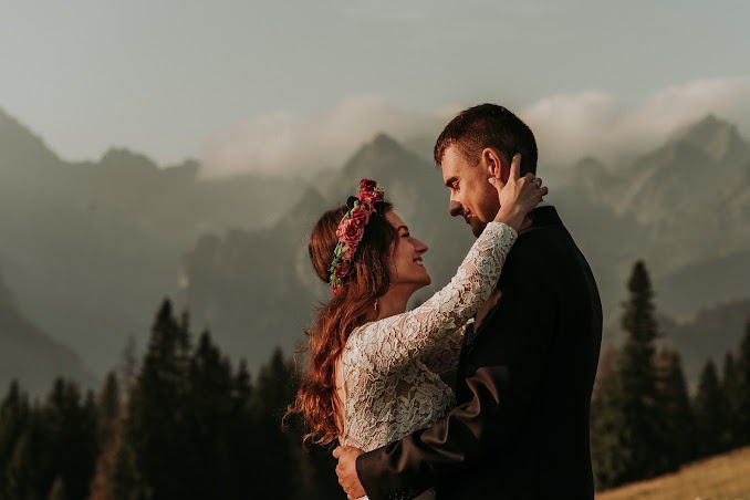 Para młoda w objęciach na tle górskim, panna młoda z wiankiem z różowych kwiatów i suknią z koronkowymi rękawami, pan młody w ciemnym garniturze, ciepłe światło zachodzącego słońca.