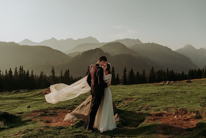 Para młoda w objęciach na tle górskiego krajobrazu, długi welon powiewa na wietrze, ciepłe światło zachodzącego słońca.