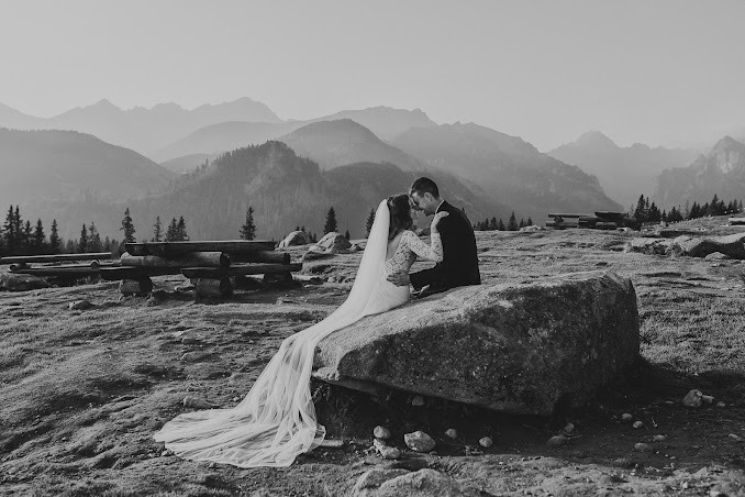 Czarno-białe zdjęcie pary młodej siedzącej na kamieniu z widokiem na góry w tle, welon panny młodej rozciąga się po kamieniu, w oddali drewniane ławki.