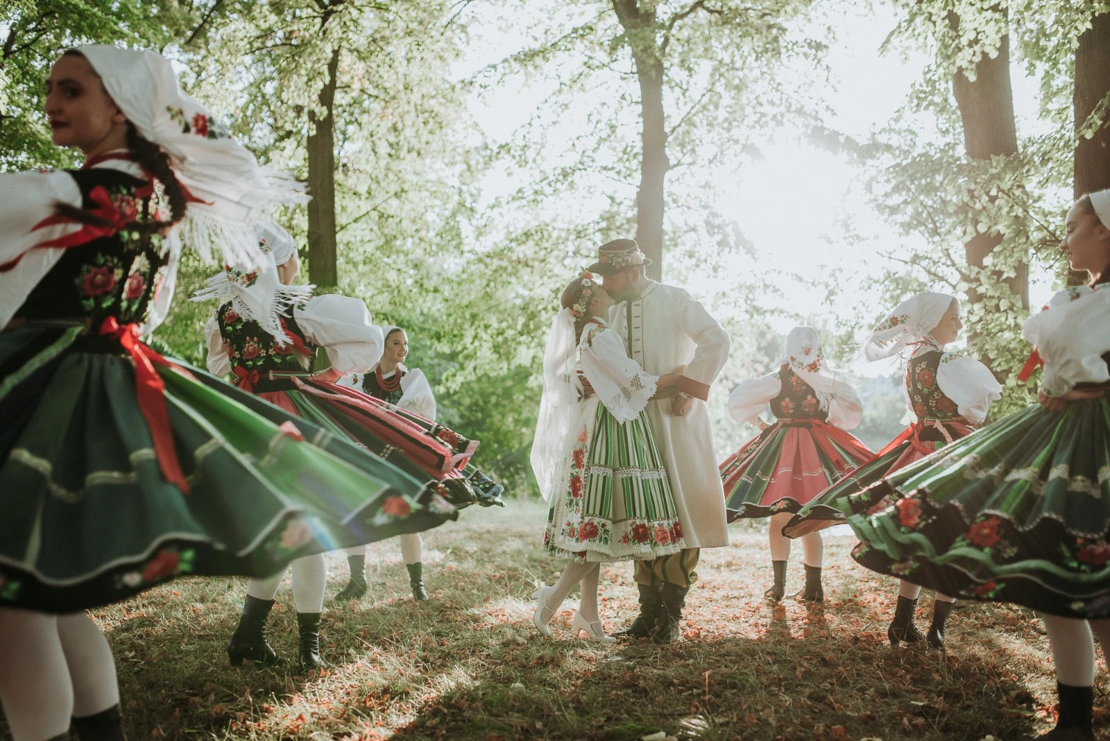 Pocałunek pary młodej w tradycyjnych strojach ludowych otoczonej przez tancerki w lesie, z promieniami słońca przebijającymi przez drzewa.