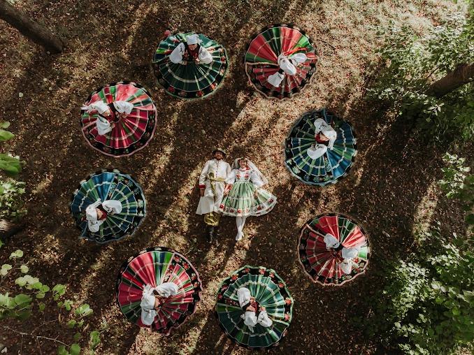 Zdjęcie z lotu ptaka przedstawia parę młodą leżącą na ziemi w otoczeniu kobiet w tradycyjnych, kolorowych strojach ludowych, tworzących okrąg wokół nich.