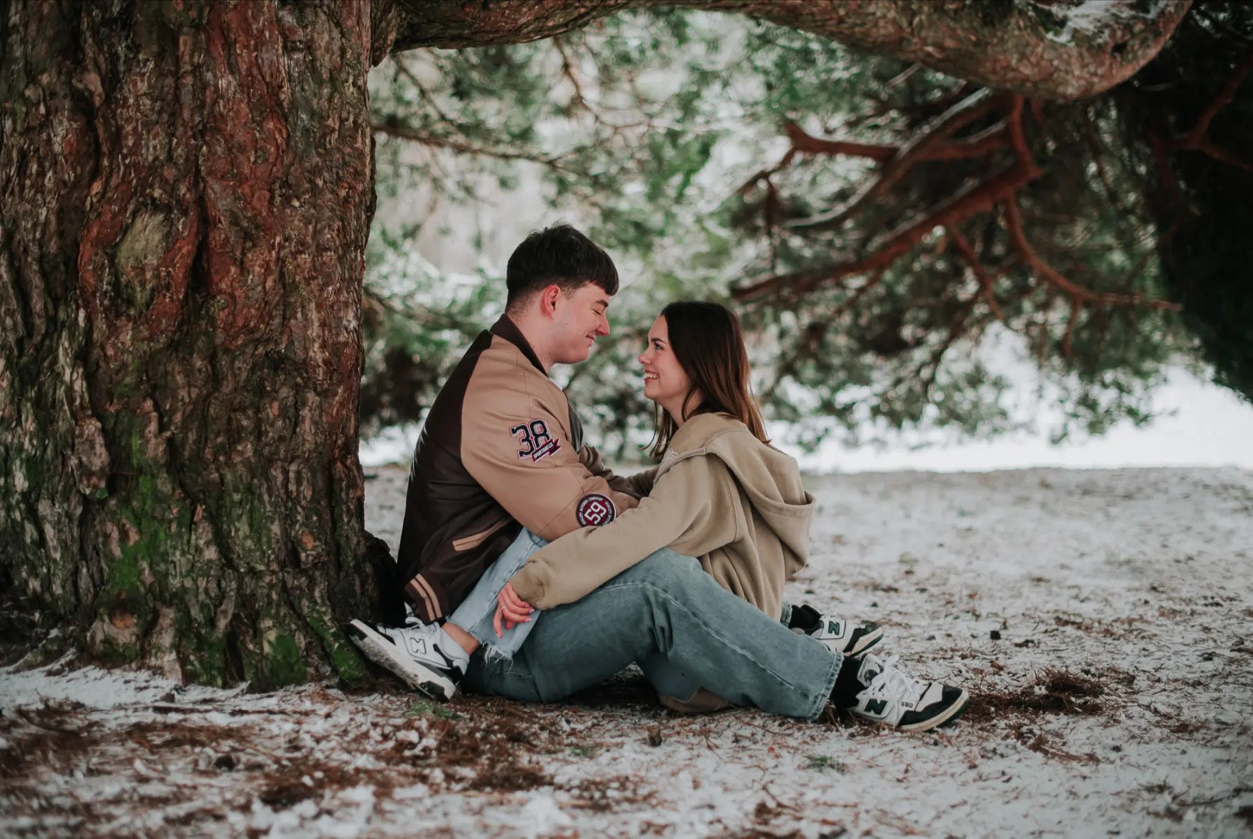 Uśmiechnięta para siedzi pod drzewem na śniegu, mężczyzna w beżowo-brązowej kurtce z numerem 38, kobieta w beżowej bluzie z kapturem, oboje w jeansach i sportowych butach.