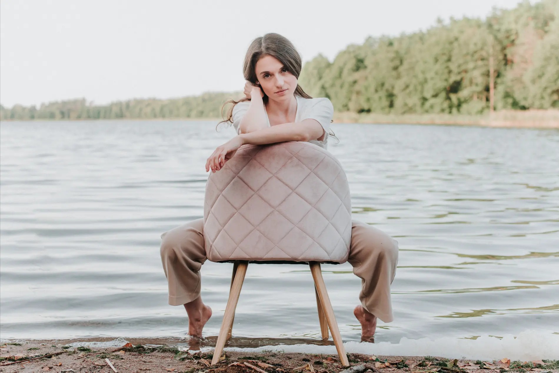 Kobieta siedzi na krześle w wodzie jeziora, patrząc w obiektyw z rękami opartymi na oparciu krzesła, w tle widok na las