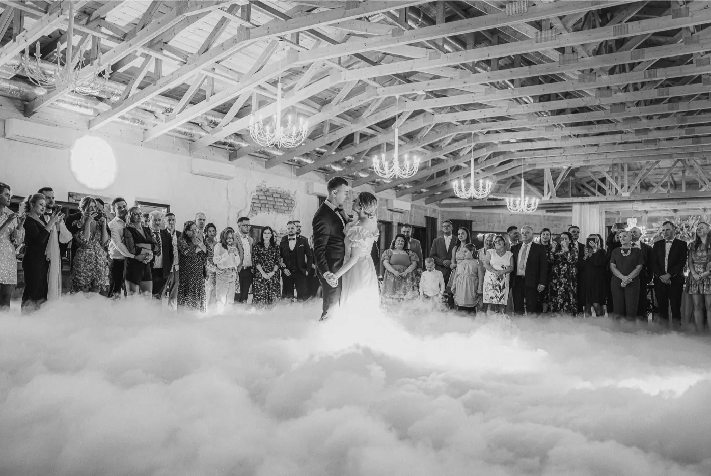 Czarno-białe zdjęcie pary młodej podczas pierwszego tańca w otoczeniu gości i efektu ciężkiego dymu, z widocznym drewnianym stropem i żyrandolami.