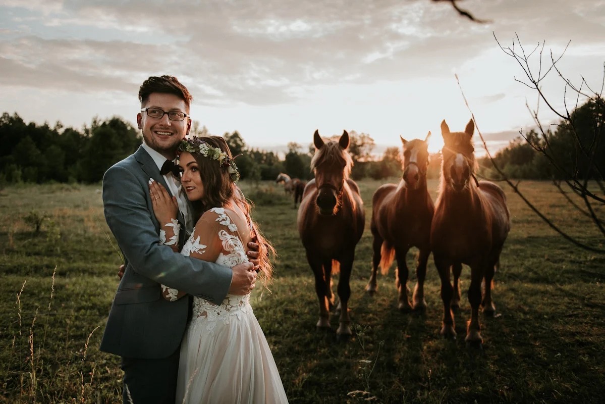 Para młoda w Ełku przytulona na łące z końmi w tle, panna młoda w sukni z koronkowymi rękawami i wiankiem, pan młody w szarym garniturze i muszce, ciepłe światło zachodzącego słońca.
