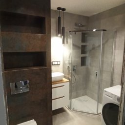 Paweł Wegner - Nowoczesna łazienka z kabiną prysznicową, umywalką nablatową na drewnianym blacie, podwieszaną toaletą i pralką LG, oświetlona podwieszanymi lampami tubami i szynoprzewodem z reflektorem.