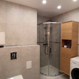 Paweł Wegner - Nowoczesna łazienka z kabiną prysznicową typu walk-in, drewnianą szafką wiszącą i pralką, wykończona w odcieniach szarości z punktowym oświetleniem sufitowym.