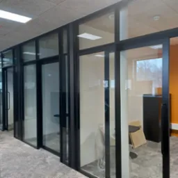 Nowoczesne biuro z przeszklonymi ścianami działowymi o czarnych ramach. Widoczne biurka, krzesła i pomarańczowa ściana w tle. Minimalistyczny design, jasne oświetlenie.