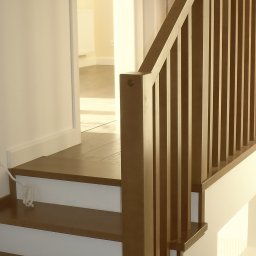Paweł Wegner - Nowoczesne schody z ciemnego drewna z białą podstopnicą i pionową balustradą, prowadzące do jasnego pomieszczenia na piętrze. Widoczny zwinięty kabel.