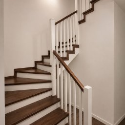 Paweł Wegner - Eleganckie, dwubiegowe schody z ciemnymi drewnianymi stopniami i białymi balustradami, oświetlone punktowym kinkietem w minimalistycznym wnętrzu.