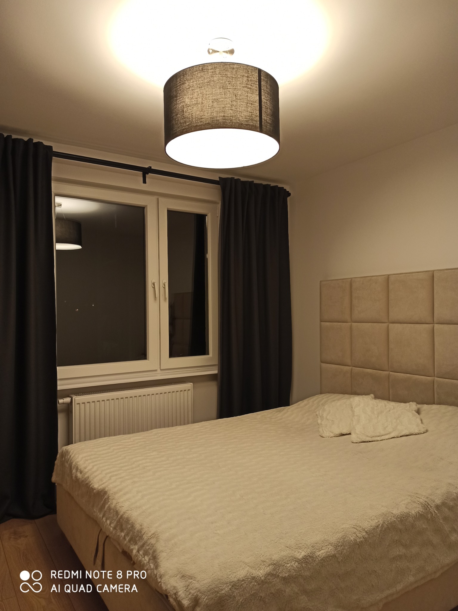 Przytulna sypialnia z beżowym łóżkiem z pikowanym zagłówkiem, oświetlona wiszącą lampą z abażurem, okno z ciemnymi zasłonami i grzejnikiem pod spodem, drewniana podłoga.