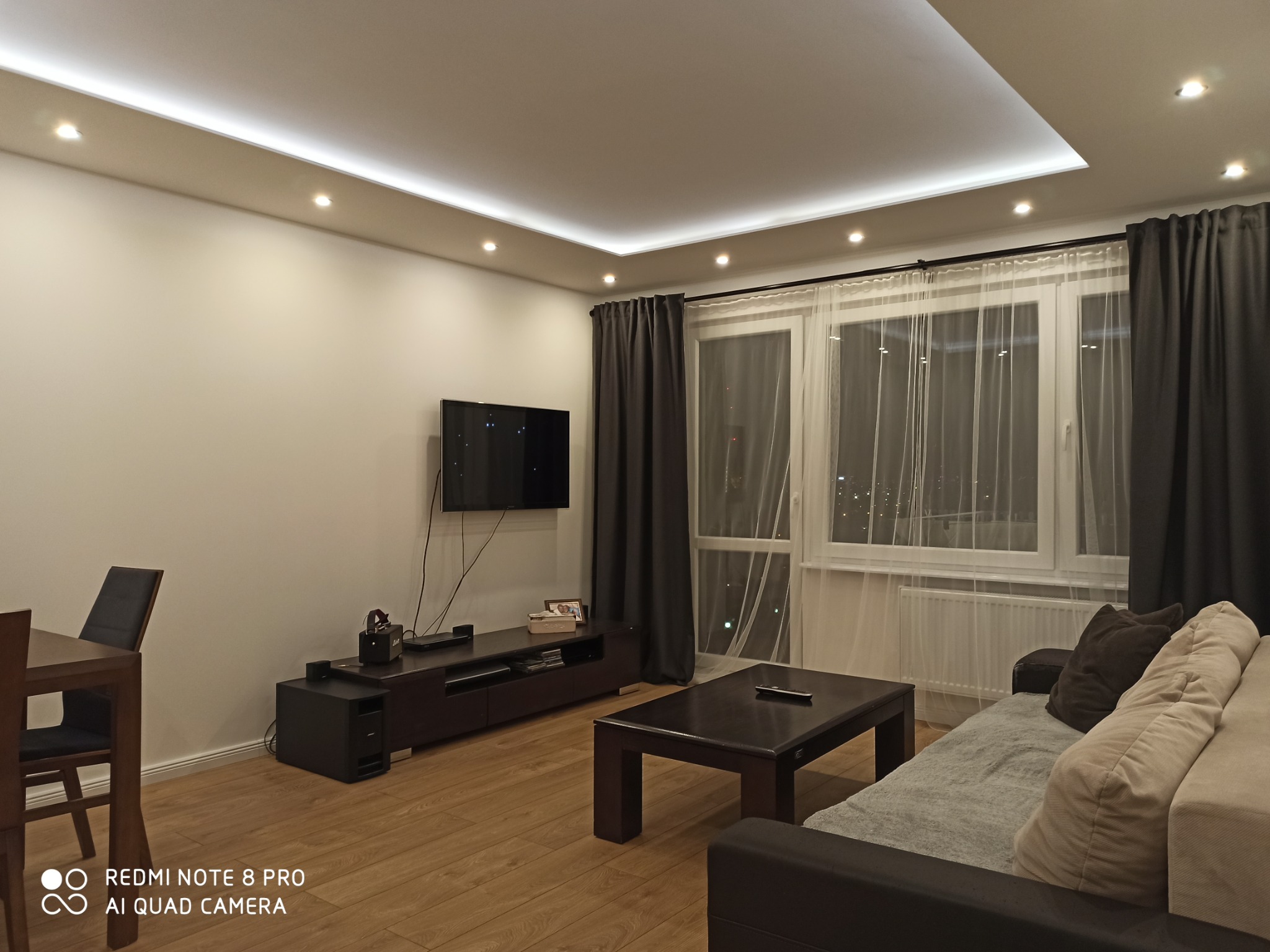 Nowoczesny salon z podwieszanym sufitem z oświetleniem LED, minimalistycznym umeblowaniem i drewnianą podłogą. Widoczne zasłony, okno z firanką i telewizor na ścianie.