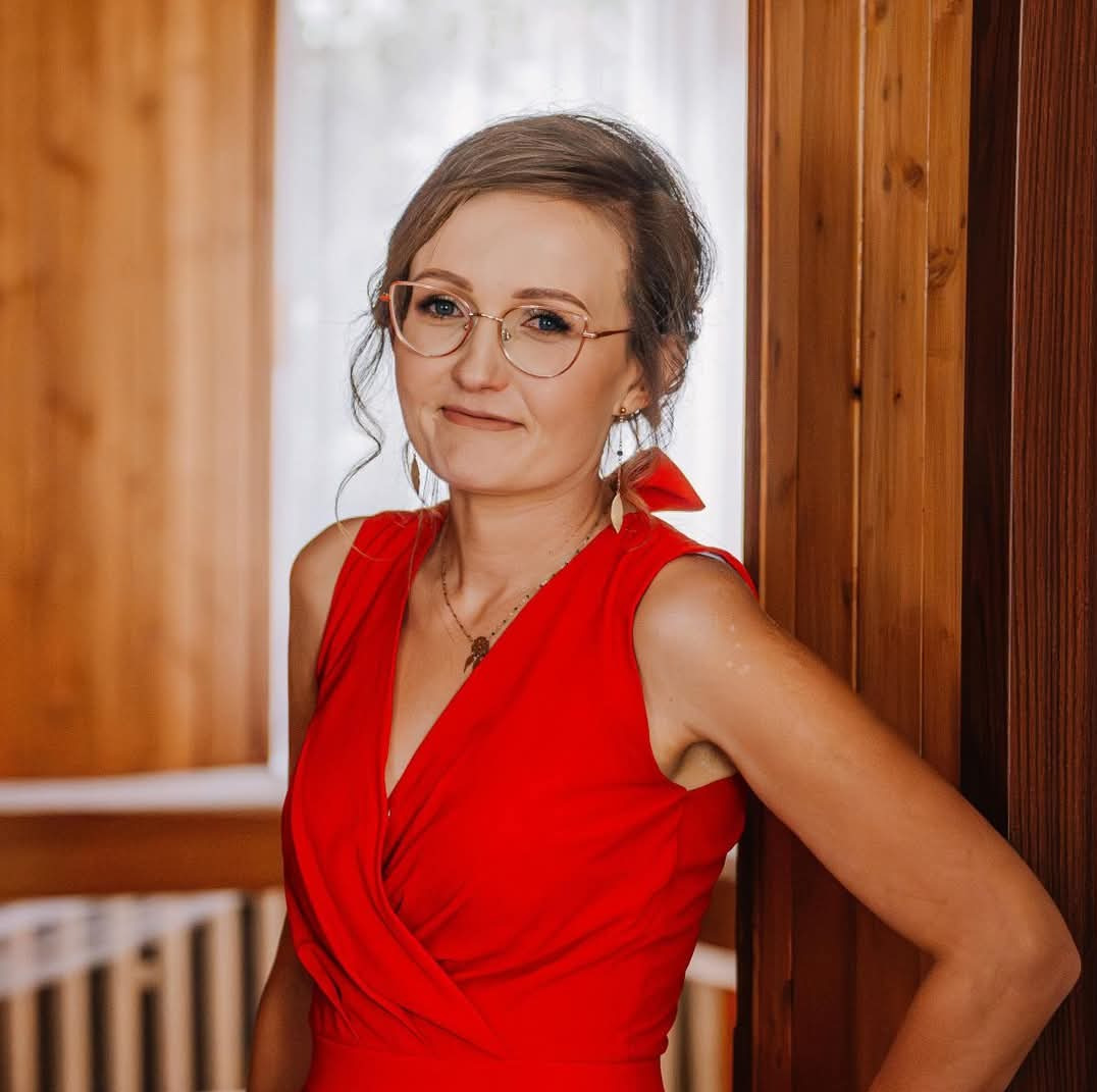 Portret uśmiechniętej kobiety w czerwonej sukience i okularach, opierającej się o drewnianą ścianę, z delikatnymi kolczykami i naszyjnikiem.