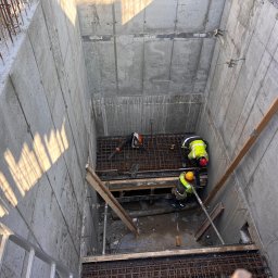 BT ŻELBET-BUD sp. z o. o - Dwóch pracowników w odblaskowych kamizelkach i kaskach układa zbrojenie wewnątrz betonowego wykopu fundamentowego, widoczna drabina i cień na ścianie.