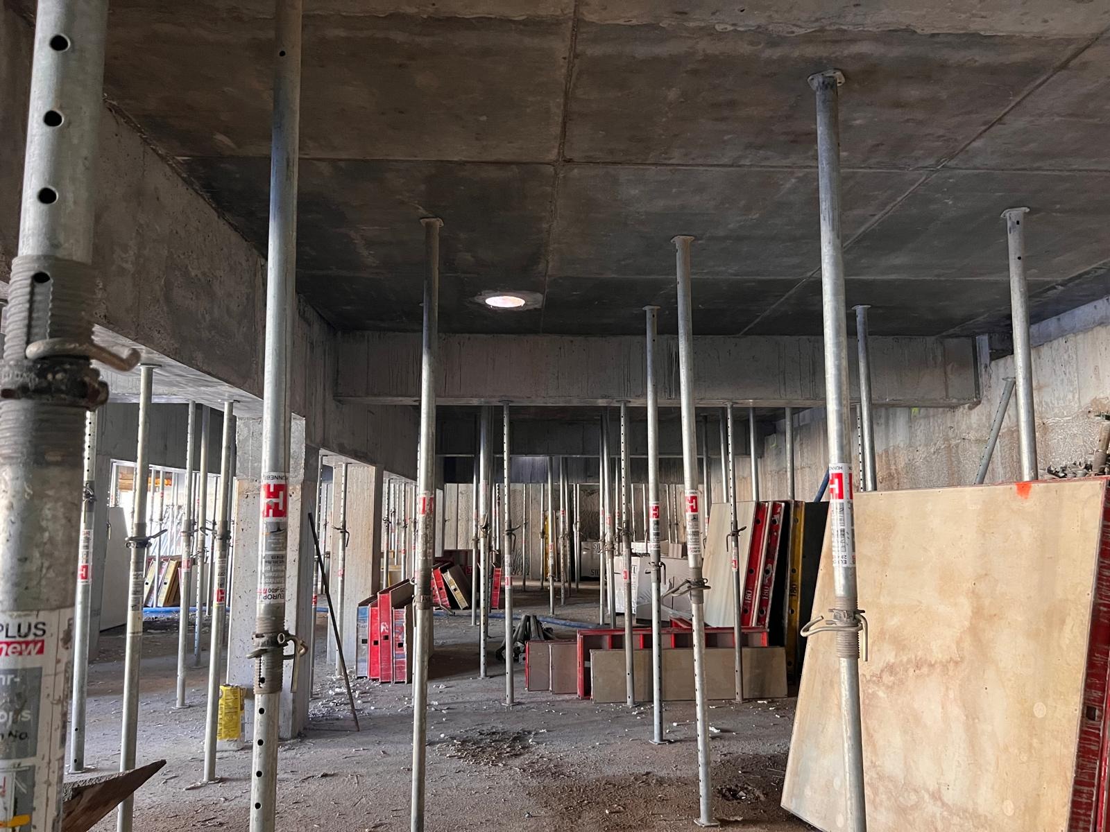 Wnętrze budynku w trakcie budowy, widoczne stalowe podpory szalunkowe podtrzymujące betonowy strop, szalunki ustawione wzdłuż ścian, surowy stan wykończenia.