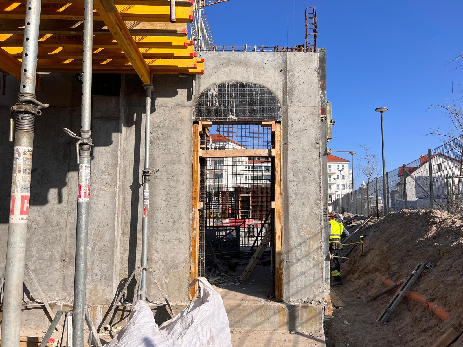 Surowa ściana z betonu z otworem na drzwi o zaokrąglonym kształcie, z widocznym zbrojeniem i deskowaniem, w tle budynki mieszkalne i pracownik w żółtej kamizelce.