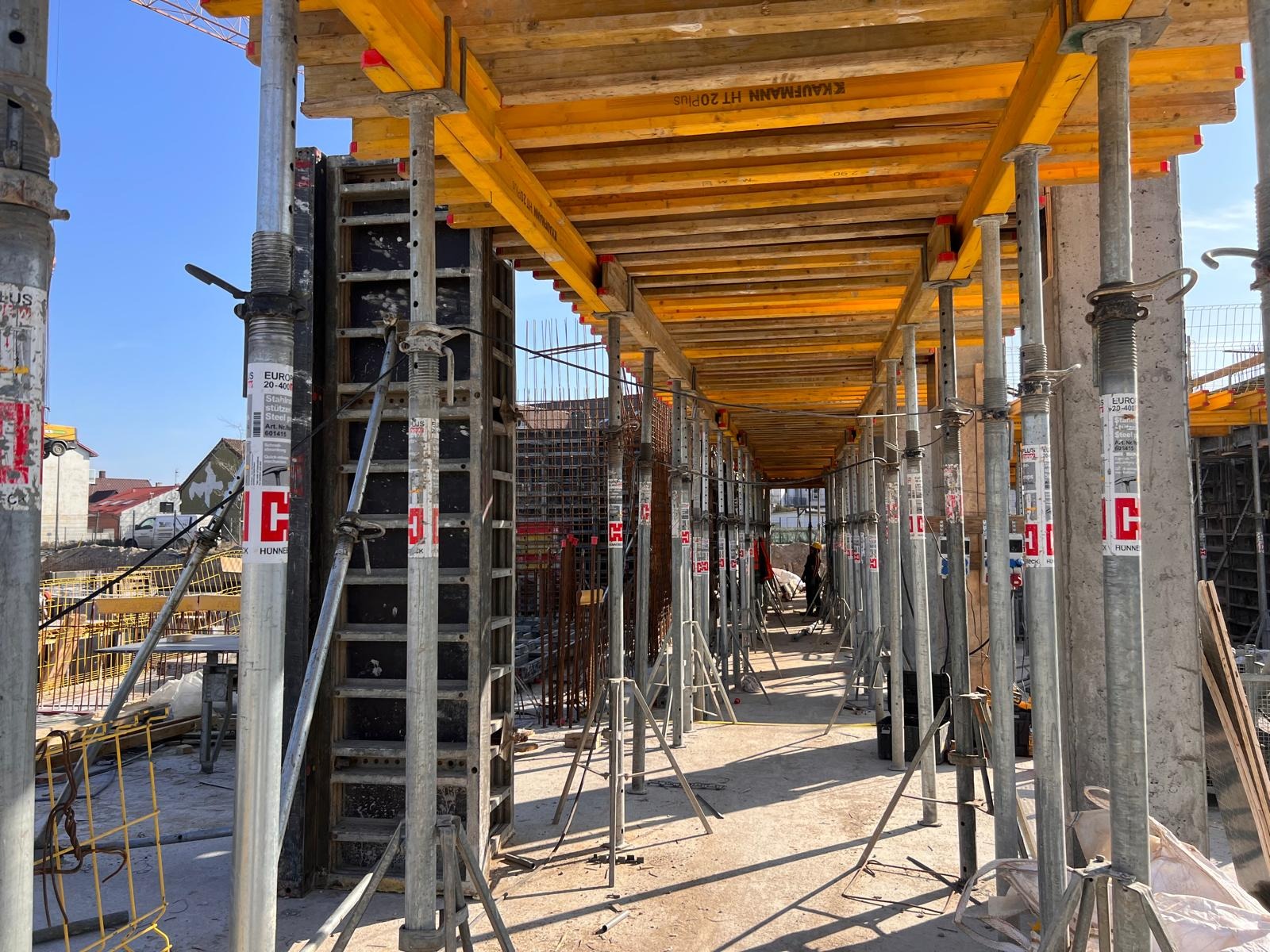 Szare, metalowe stemple budowlane podtrzymujące żółte, drewniane szalunki stropowe na placu budowy w słoneczny dzień. W tle widoczne zbrojenia i fragment zabudowy mieszkalnej.
