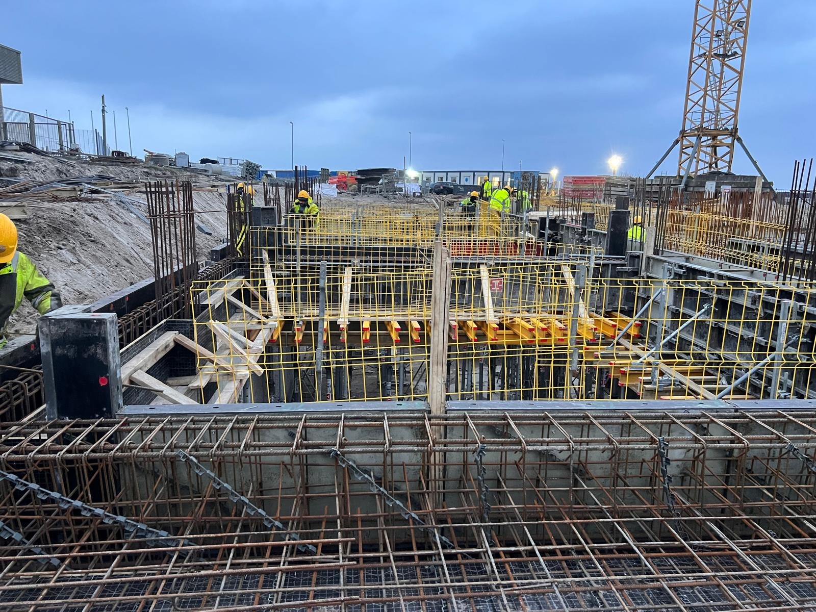 Zbrojenie fundamentów z żółtymi elementami szalunkowymi na budowie w pochmurny dzień, widoczni pracownicy w odblaskowych ubraniach i dźwig w tle.